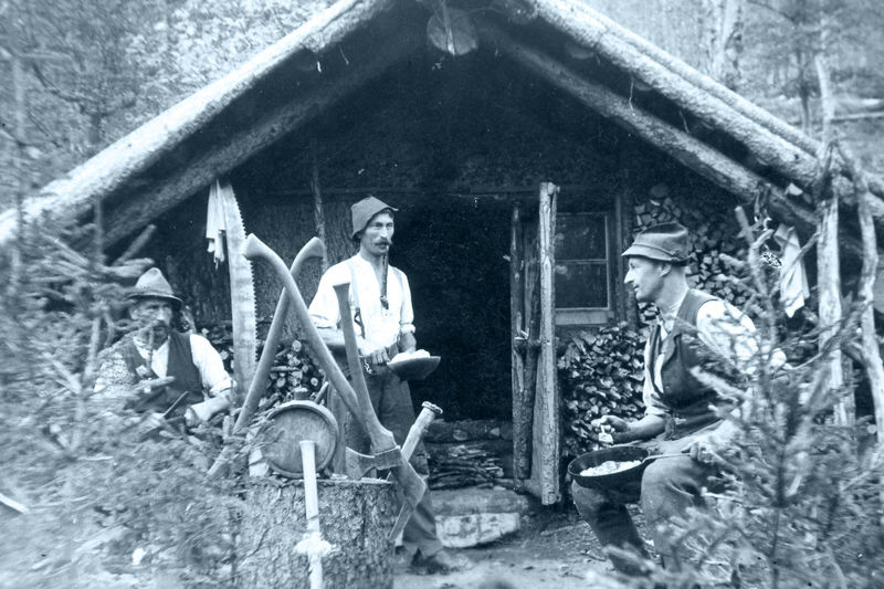 Historische schwarz-weiß Aufnahme; drei Holzknechte essen vor Hütte